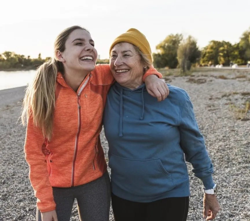 A granddaughter and grandmother wearing jackets smile and walk together alongside a river