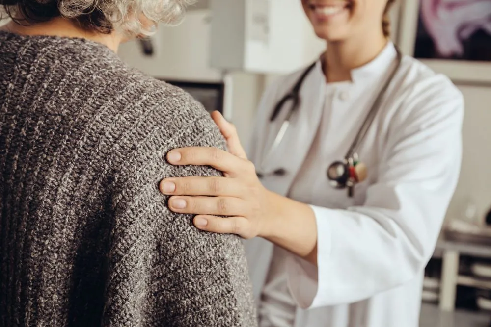 A female doctor in a white lab coat rests a comforting hand on a patient's shoulder