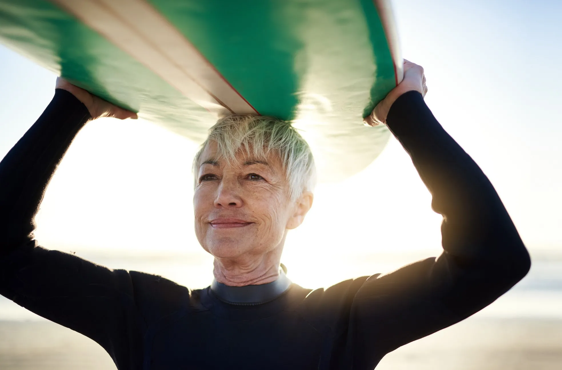 A senior woman with short gray hair holds a surf board over her head on a sunny beach