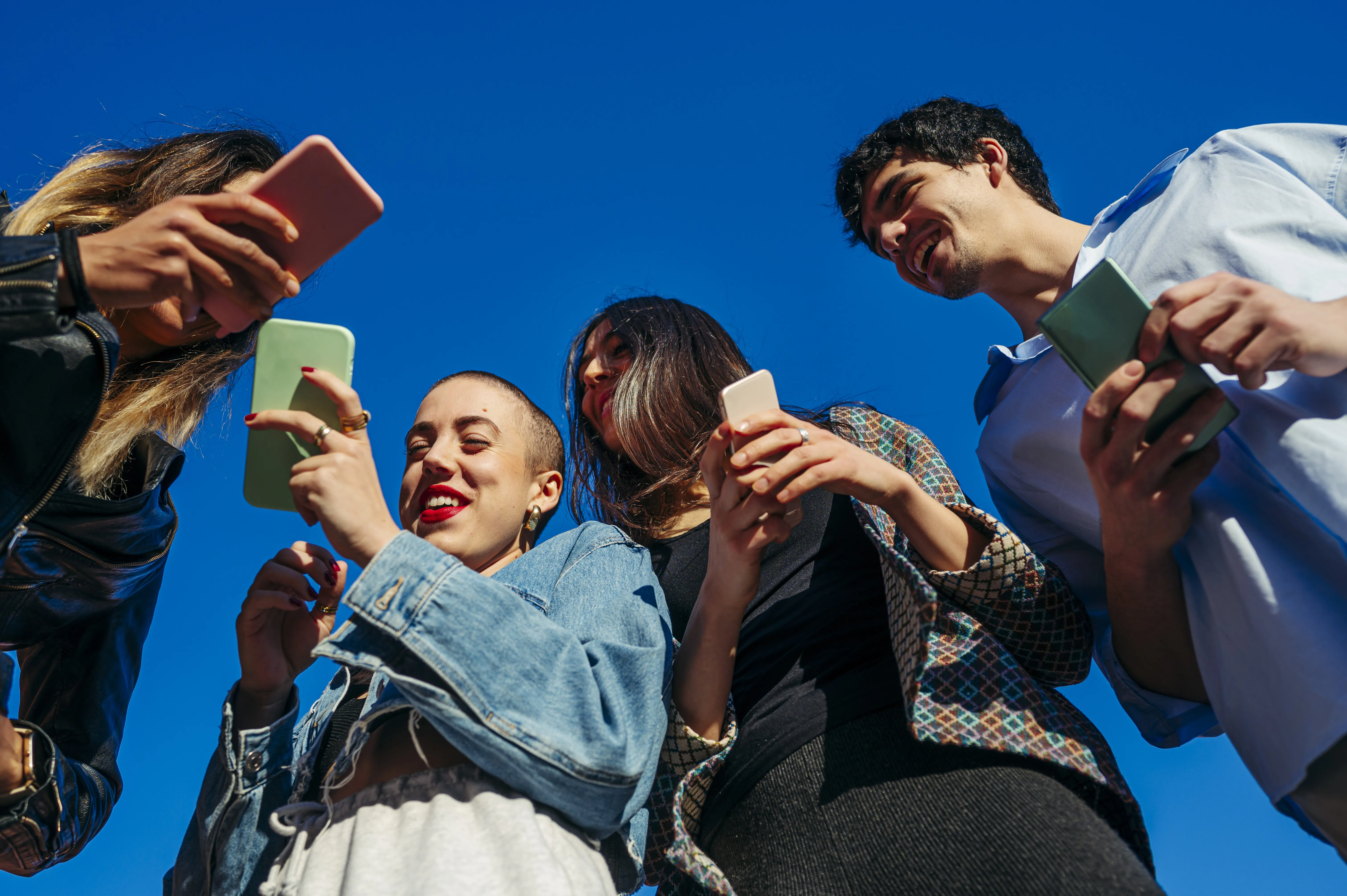 A group of smiling young adults stand together against a blue sky while looking at their smartphones