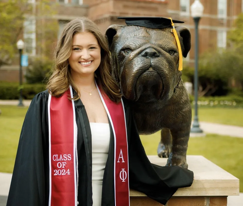 Abbie smiles in a black graduation gown, standing outside on her college campus