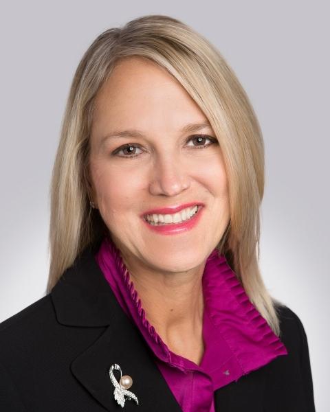 A headshot of CSC's new CEO Debbie Weir shows her smiling and wearing a purple blouse and black blazer with a cancer awareness ribbon lapel pin