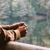 An unseen person rests their hands on wood bridge looking out at a reflective tree-lined lake