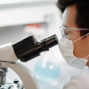 A lab technician wearing a mask looks through a microscope