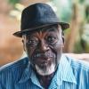 An African American elderly man wearing a hat smiles from his seat on his porch