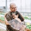 A man hugs his wife as they stand inside a sunlit greenhouse