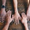 People of diverse races and genders rest their hands beside one another on a wooden table