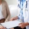 A doctor in a white coat holds a clipboard while talking with a patient