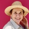 An elderly woman wearing a white blouse and a sun hat smiles in front of a bright pink wall
