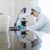 A female lab technician wearing a white coat and cap peers into a microscope positioned on a reflective tabletop