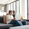 A happy couple sits together on their sunlit living room couch looking at their laptop