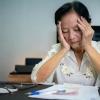 A distressed elderly woman rests her face in her hands as she looks at some bills on her desk