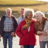 A group of smiling senior friends wearing scarves and jackets walks along a breezy shoreline