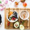 A breakfast displays fresh fruits sits alongside a wood cutting board with a bowl of cereal, avocado, and a cup of tea