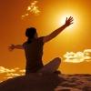 A woman sitting on a rock extends her arms out as if embracing the setting sun in an orange-lit sky