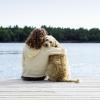 A young woman hugs her dog as they sit together on the edge of a pier overlooking a lake