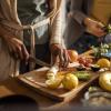 Women prepare veggies and fruits on a cutting board in a brightly lit kitchen