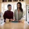 A mother and daughter smile while looking at a digital tablet on their kitchen table