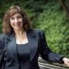 Kerri Susko wears a black business suit and smiles from a wooden bridge beside trees