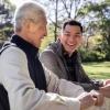 A senior man in a jacket vest laughs with his son outdoors where they rest their hands on a fence