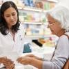 A senior woman stands at a pharmacy counter showing a female pharmacist her prescription