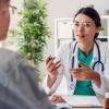 A woman doctor in a white lab coat speaks with a man sitting at a desk in a doctor's office