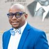 Tony Walker wears a blue and white bow tie, blue blazer, white blouse, and glasses with blue frames
