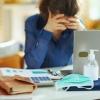 A woman in a blue blouse rests her face in her hands at a desk with a laptop, calculator, and worksheets