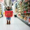 A woman carrying a red grocery basket stands in the middle of a supermarket aisle
