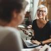 A woman with short gray hair and dark glasses smiles with a phone and laptop while talking with an unseen woman