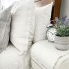 Pillows are stacked on a bed next to a table with alarm clock and potted lavender flowers