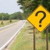 A yellow road sign on the side of an empty road displays a large question mark