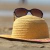 A pair of brown sunglasses sits on a straw sun hat resting on a sandy beach