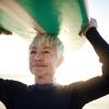 A senior woman with short gray hair holds a surf board over her head on a sunny beach