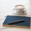 A black pen rests on a hard-cover notebook beside a cup of coffee
