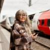 A mature woman with long hair and dark glasses waits at a train station