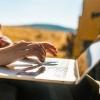 Unseen person wearing a flannel shirt types on a laptop outdoors in a mountain landscape