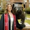 Abbie smiles in a black graduation gown, standing outside on her college campus