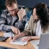A man and a woman seated at a counter with a calculator discuss paperwork